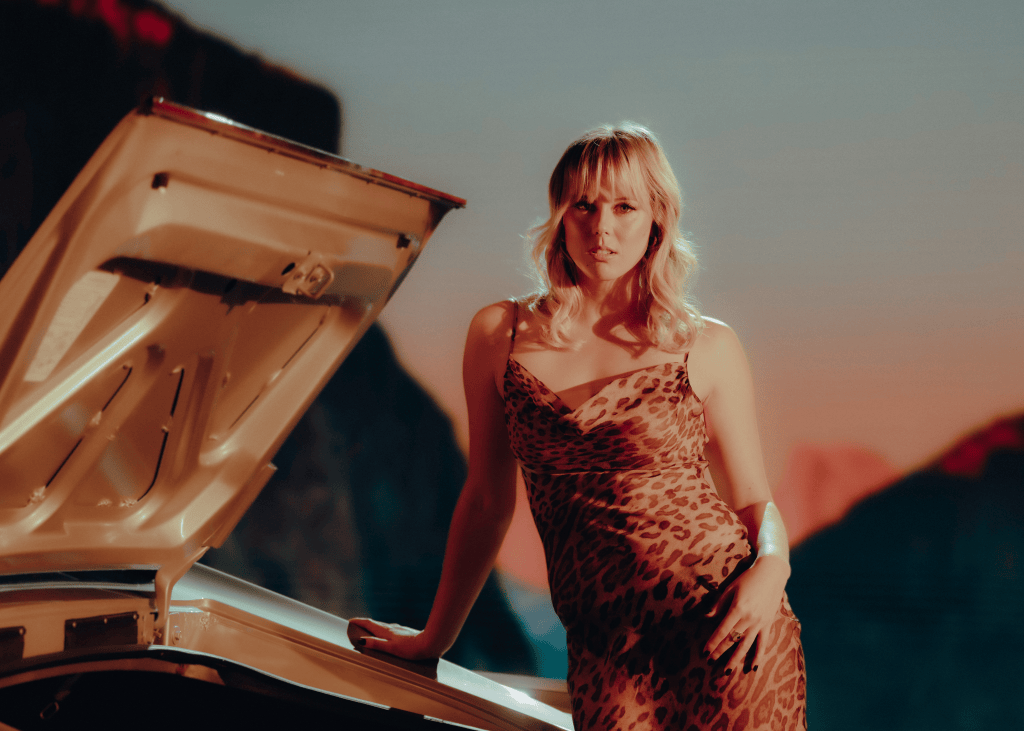 A woman in a leopard print dress leans against the open boot of a car, with a scenic landscape and sunset in the background.