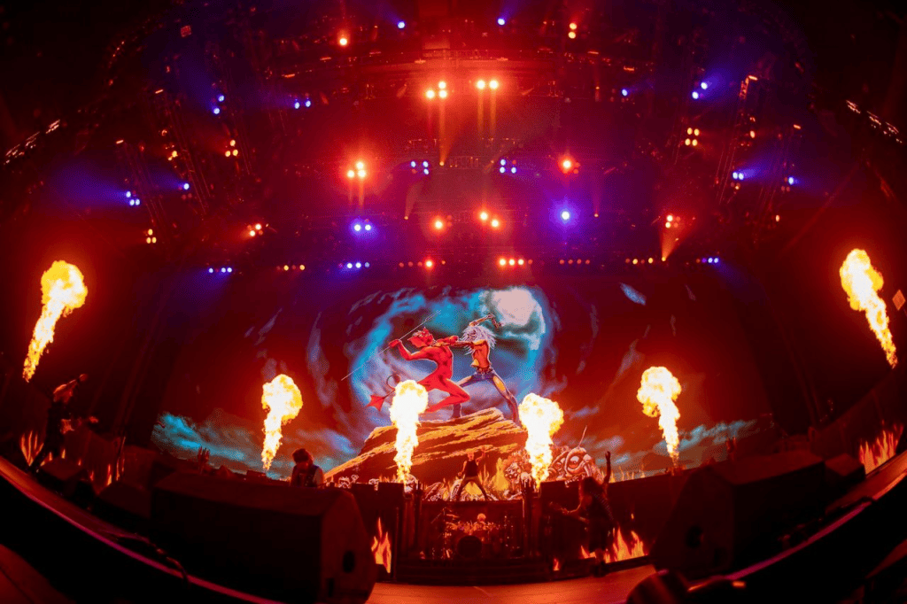 A vibrant concert stage featuring dramatic lighting and pyrotechnics, with a large backdrop displaying a fierce battle scene.