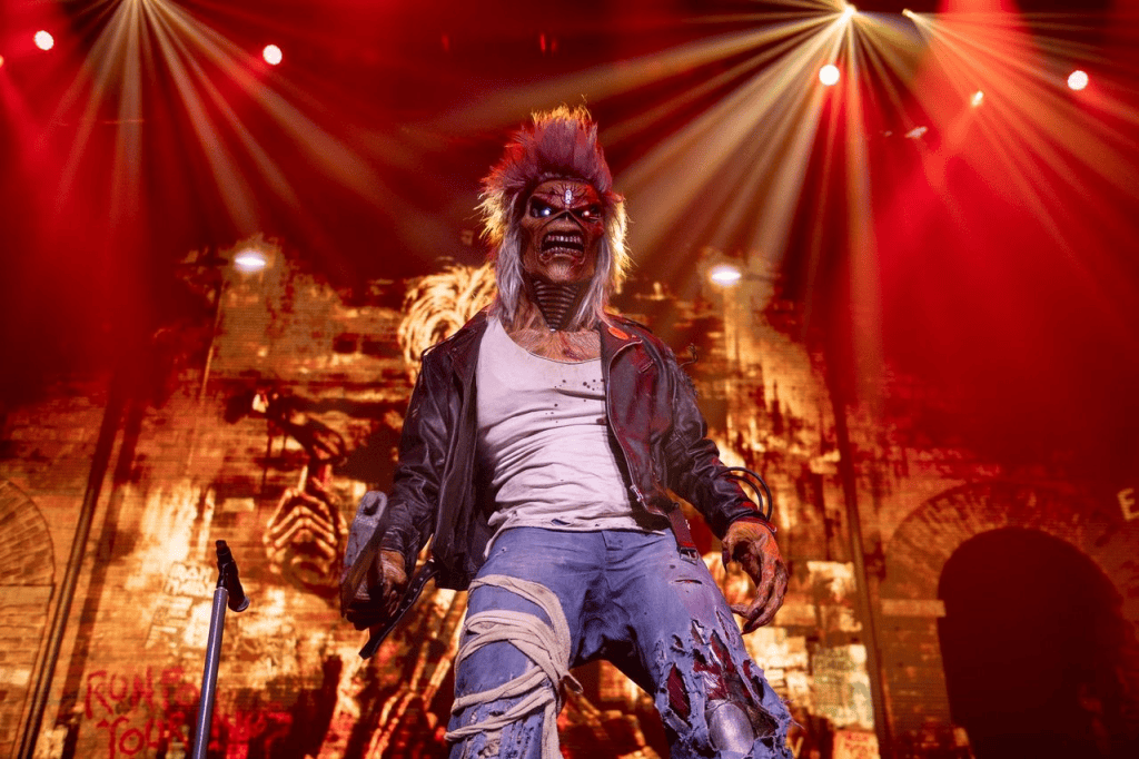 A performer dressed as a monster with wild hair and a leather jacket, standing on stage with dramatic lighting.