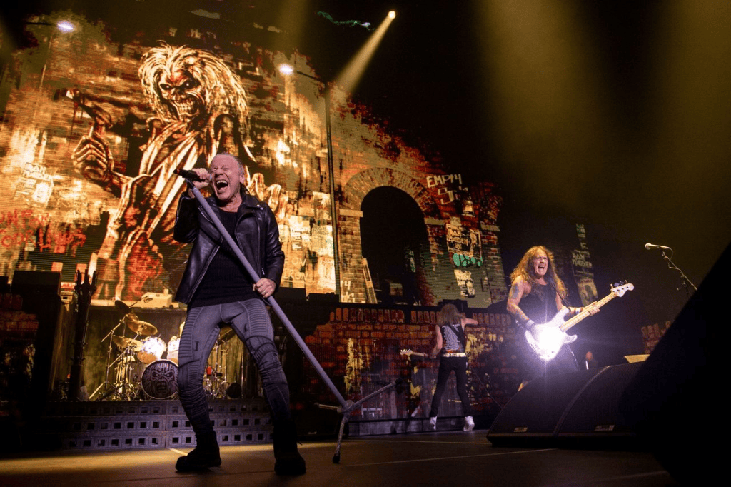 A rock band performs on stage with dramatic lighting and a large backdrop featuring an iconic character. The lead singer, wearing a leather jacket, passionately sings into a microphone while holding a microphone stand. A guitarist plays beside him.